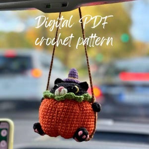 May include: A crocheted black cat wearing a purple witch hat is inside an orange pumpkin. The cat is hanging from the rearview mirror of a car. The text "Digital PDF crochet pattern" is above the cat.