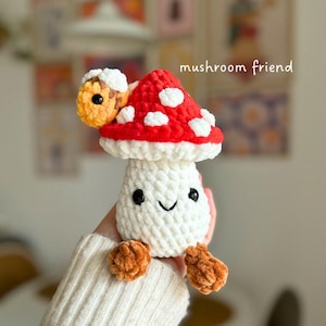 May include: A handmade crochet mushroom toy with a red cap and white spots, a white stem, and orange feet. The mushroom has a smiling face with black eyes. The text "mushroom friend" is written in white.
