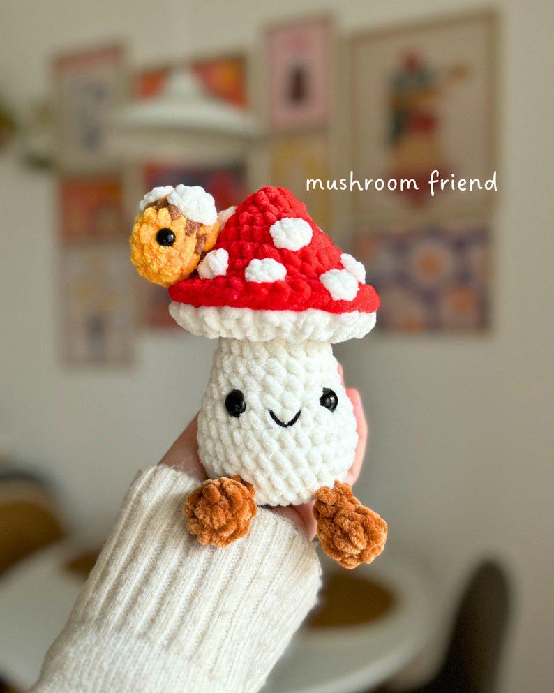 May include: A handmade crochet mushroom toy with a red cap and white spots, a white stem, and orange feet. The mushroom has a smiling face with black eyes. The text "mushroom friend" is written in white.