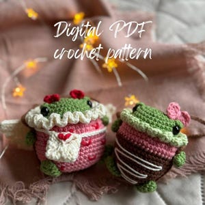 May include: Two crocheted frog amigurumi toys. One is pink and white with a heart and the other is brown and pink with white stripes. The toys are on a pink and white patterned fabric. Digital PDF crochet pattern.