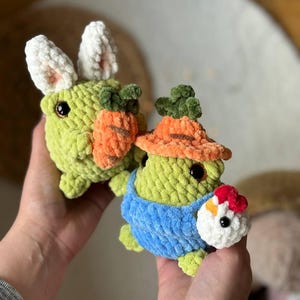 Pu&ograve; includere: Due giocattoli di peluche all'uncinetto, uno un coniglio verde con orecchie bianche che tiene una carota e l'altro una rana verde che indossa una salopette blu e un pollo all'uncinetto rosso e bianco.