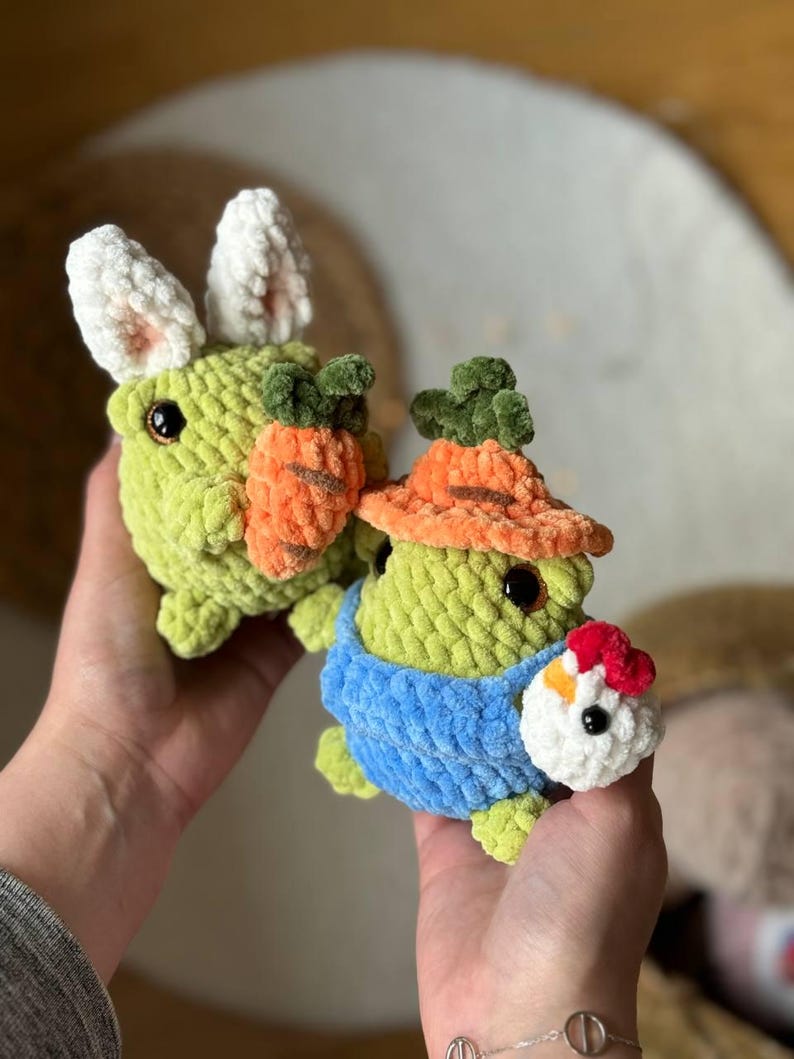 Pu&ograve; includere: Due giocattoli di peluche all'uncinetto, uno un coniglio verde con orecchie bianche che tiene una carota e l'altro una rana verde che indossa una salopette blu e un pollo all'uncinetto rosso e bianco.
