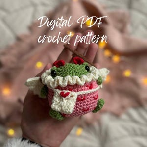 May include: A crocheted amigurumi toy in the shape of a frog wearing a pink and white dress with hearts. The frog has green skin, black eyes, and a red bow. The toy is being held in a hand. The text "Digital PDF crochet pattern" is visible above the toy.