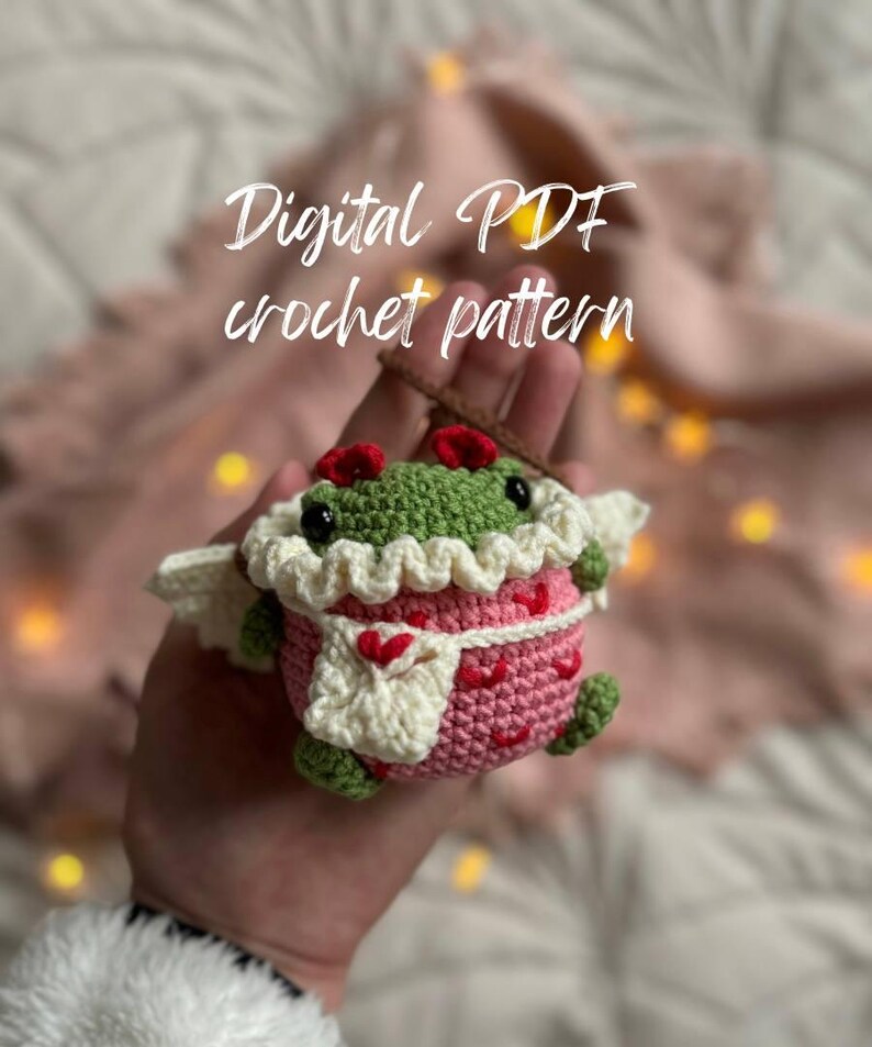 May include: A crocheted amigurumi toy in the shape of a frog wearing a pink and white dress with hearts. The frog has green skin, black eyes, and a red bow. The toy is being held in a hand. The text "Digital PDF crochet pattern" is visible above the toy.