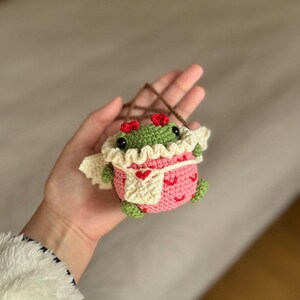 May include: A small crocheted frog wearing a pink and white dress with red hearts. The frog has green skin and black eyes.