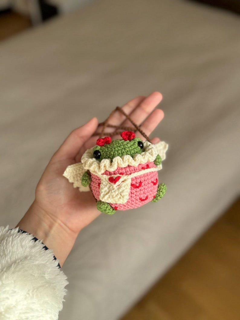 May include: A small crocheted frog wearing a pink and white dress with red hearts. The frog has green skin and black eyes.