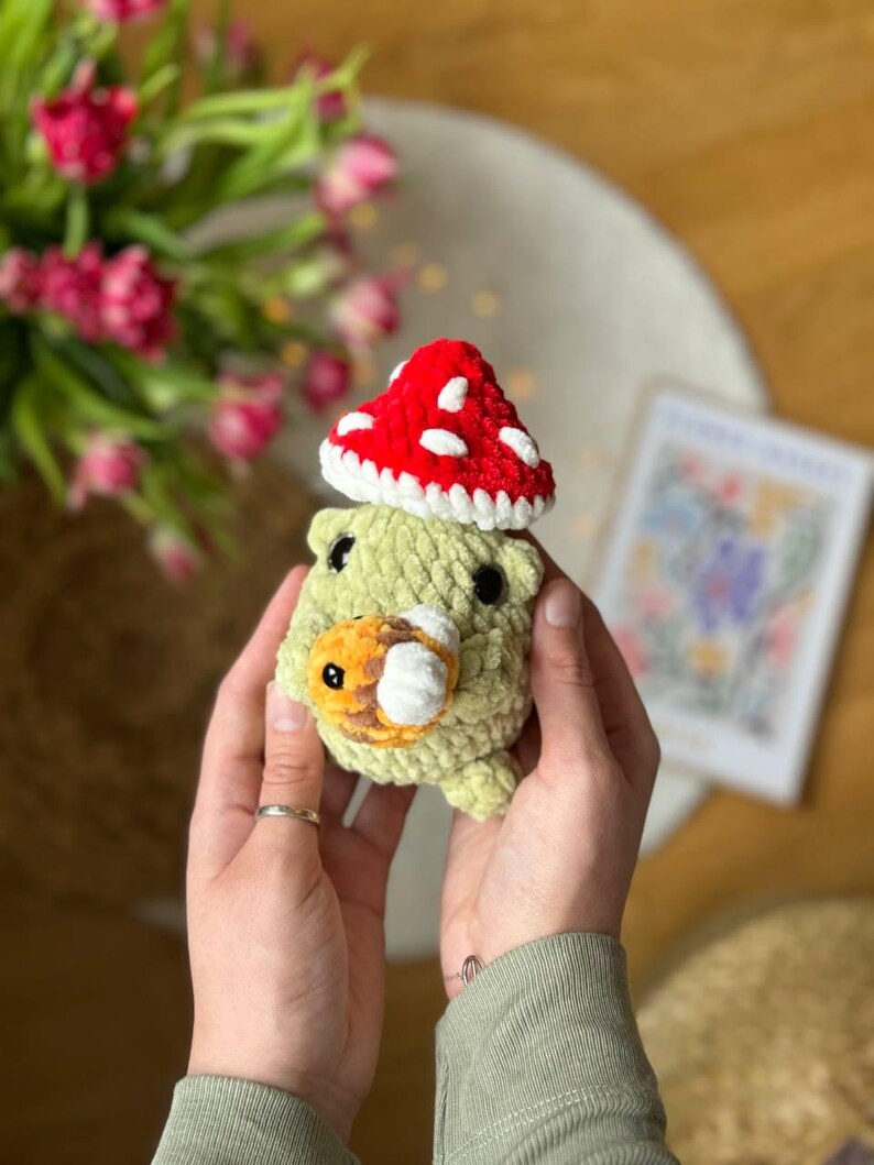 Puede incluir: Un animal de peluche tejido a crochet con forma de hongo, con un sombrero rojo y blanco y un cuerpo verde. El hongo est&aacute; siendo sostenido en las manos de una persona.