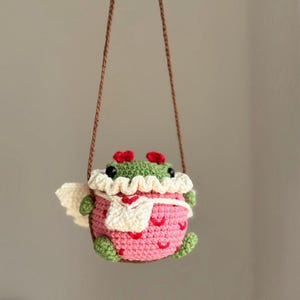 May include: A crocheted frog wearing a pink and white dress with a white scarf and red accents. The frog is hanging from a brown string.