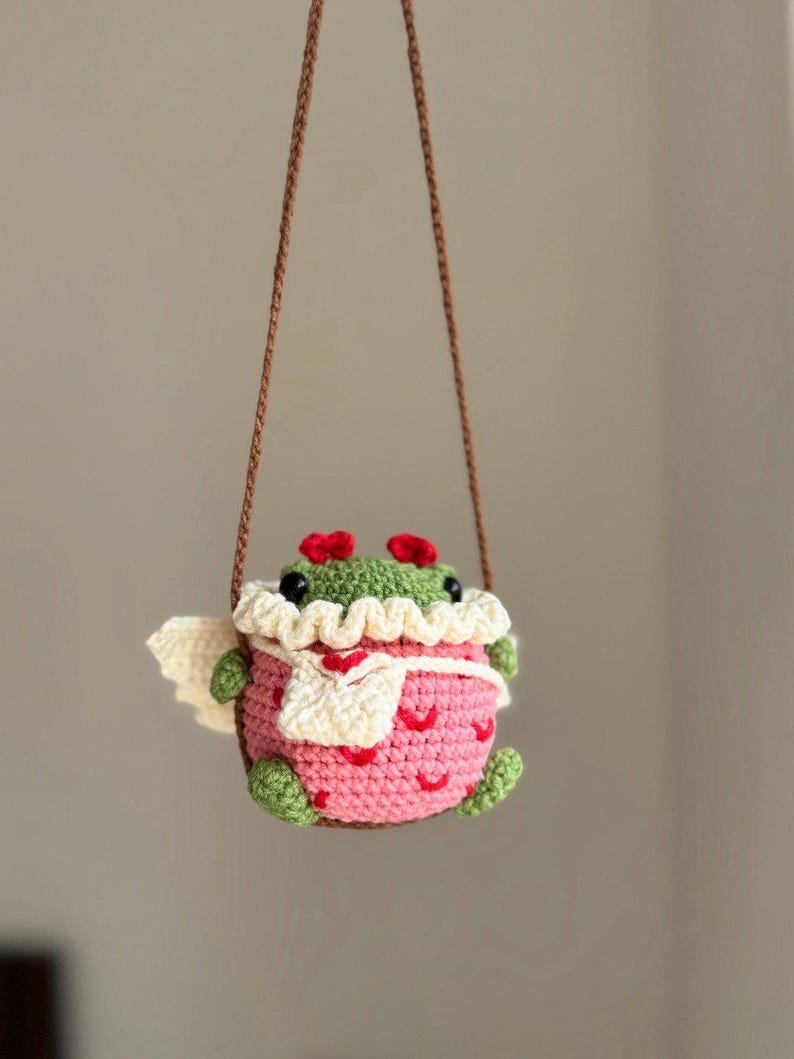 May include: A crocheted frog wearing a pink and white dress with a white scarf and red accents. The frog is hanging from a brown string.
