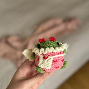 May include: A crocheted green frog wearing a pink and white dress with a heart pattern. The frog has red hearts for ears and is holding a white crocheted heart.