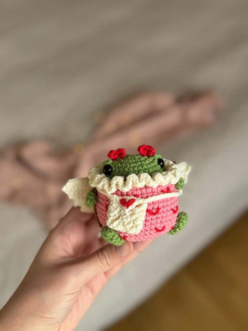 May include: A crocheted green frog wearing a pink and white dress with a heart pattern. The frog has red hearts for ears and is holding a white crocheted heart.