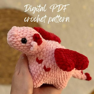 May include: A pink crocheted dragon with red wings. The dragon has a small, black nose and eyes. The text "Digital PDF crochet pattern" is written above the dragon.