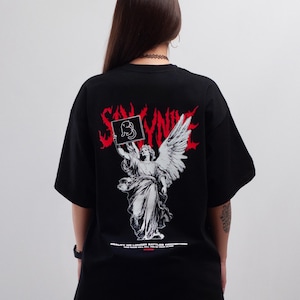 Puede incluir: Una camiseta negra con un gráfico grande en la espalda. El diseño incluye una ilustración blanca de un ángel sosteniendo un cartel con un dibujo de un gato, texto rojo estilizado arriba y el texto blanco "REALITY NO LONGER BATTLES PERCEPTION" abajo.