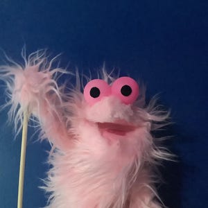 May include: A pink puppet with large, round, pink eyes and a wide, smiling mouth. The puppet has fluffy, shaggy fur against a blue backdrop. A wooden stick is visible, likely used for manipulation.