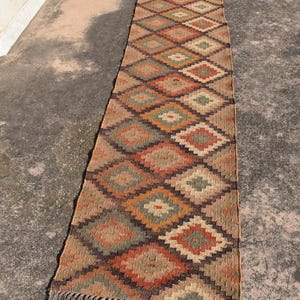 Puede incluir: Una alfombra larga y rectangular con un patrón de diamantes repetido en tonos de marrón, naranja, verde y crema. La alfombra tiene un borde con flecos y está hecha de un material tejido. La alfombra está colocada sobre una superficie de hormigón.