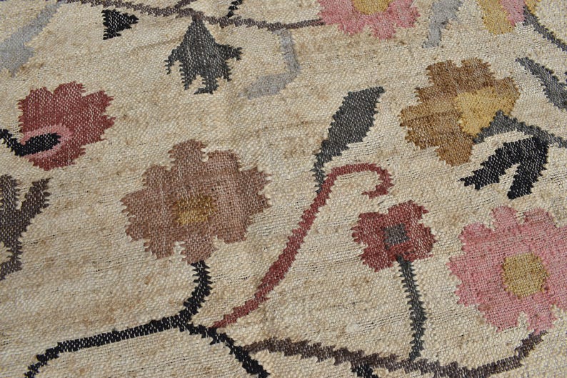 May include: Close-up of a beige rug with a floral pattern. The rug features flowers in shades of pink, brown, and yellow, with black stems and gray leaves. The rug has a woven texture.