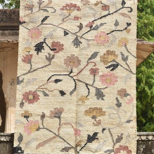May include: A rectangular beige rug with a floral pattern. The rug features pink, brown, yellow, and black flowers with gray and brown stems and leaves. The rug has a fringed edge.