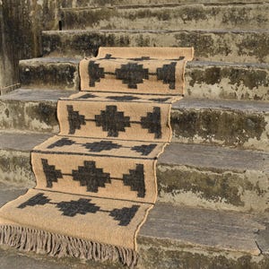 Op de afbeelding: Een tan en zwarte geometrische jute loper met franjes aan de uiteinden. De loper ligt op een set betonnen treden.
