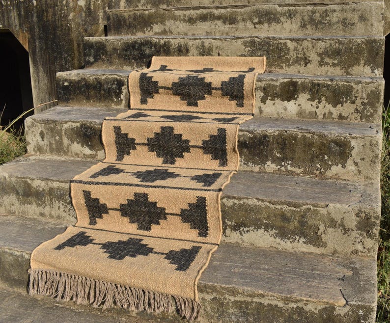 Op de afbeelding: Een tan en zwarte geometrische jute loper met franjes aan de uiteinden. De loper ligt op een set betonnen treden.