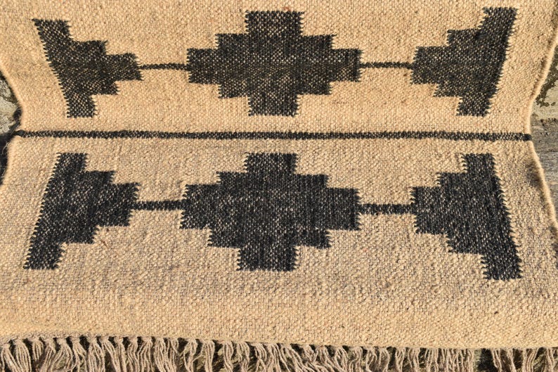 Op de afbeelding: Een geweven tapijt in tan en zwart met een geometrisch patroon. Het tapijt heeft een franje rand.