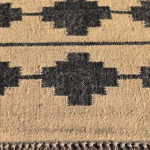 Op de afbeelding: Een geweven beige en zwart tapijt met een geometrisch patroon. Het tapijt heeft een franje rand.