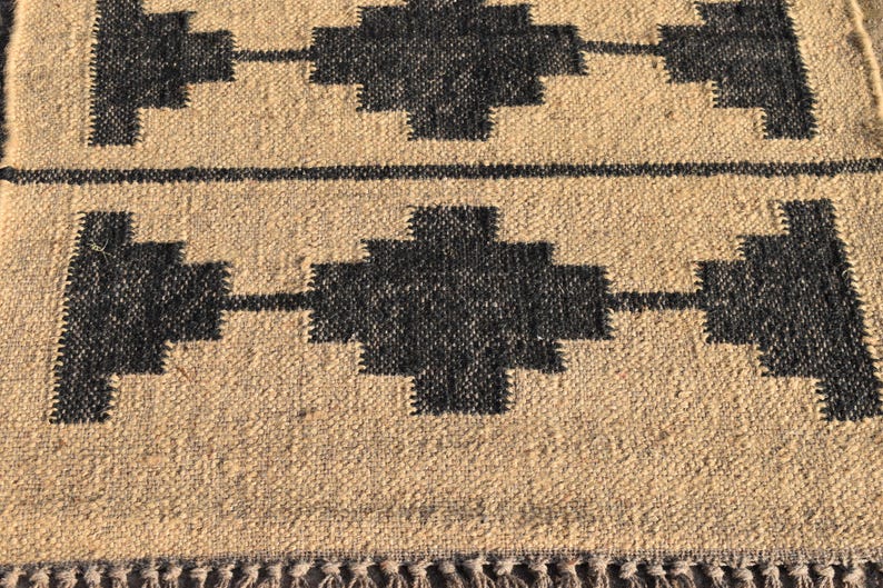 Op de afbeelding: Een geweven beige en zwart tapijt met een geometrisch patroon. Het tapijt heeft een franje rand.