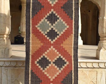 May include: A long, rectangular rug with a geometric pattern in shades of red, black, beige, and green. The rug has a fringe border.
