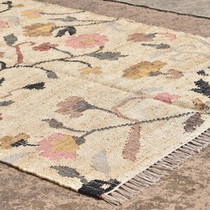 May include: A rectangular beige rug with a floral pattern. The rug features pink, yellow, and brown flowers with black stems and gray leaves. The rug has a fringed edge and is made of a woven material.