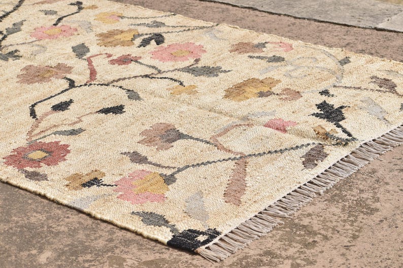 May include: A rectangular beige rug with a floral pattern. The rug features pink, yellow, and brown flowers with black stems and gray leaves. The rug has a fringed edge and is made of a woven material.