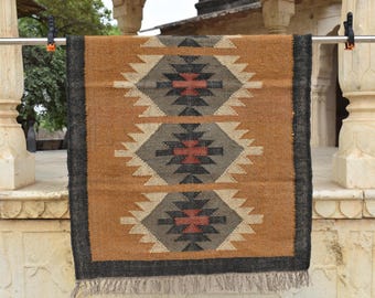 May include: A rectangular rug with a geometric pattern. The rug features a tan background with a black border and repeating diamond shapes in gray, white, and red. The rug is made of a woven material and has a fringed edge.