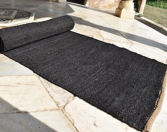 May include: A rolled-up black jute rug with a woven texture. The rug is partially unrolled, revealing its rectangular shape and a natural fiber border. The rug is placed on a light-colored stone surface.