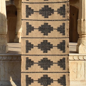 Op de afbeelding: Een geometrisch gestreept tapijt in zwart en beige met franjes aan de uiteinden. Het tapijt is gemaakt van natuurlijke vezels en heeft een rustieke uitstraling.
