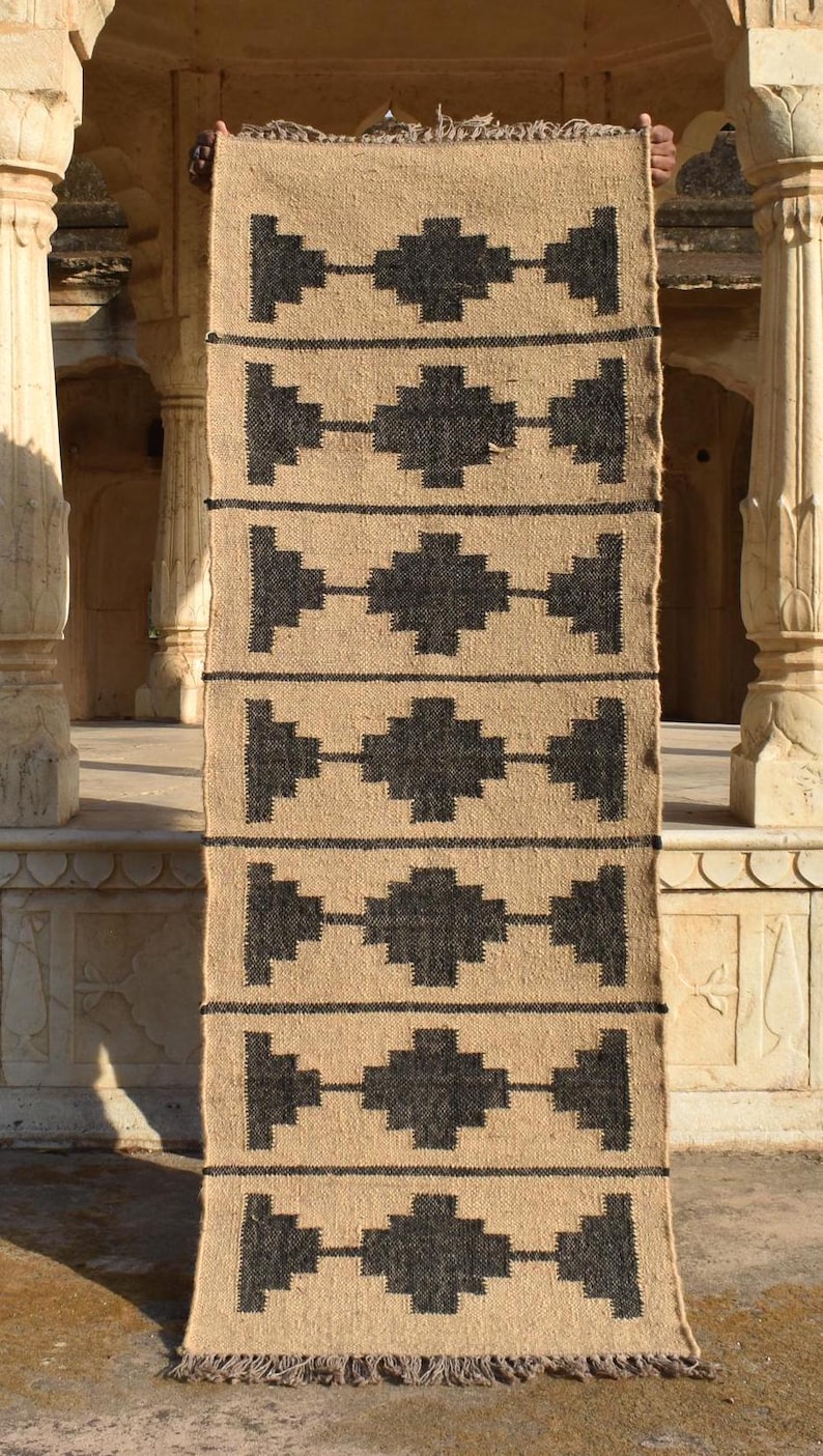 Op de afbeelding: Een geometrisch gestreept tapijt in zwart en beige met franjes aan de uiteinden. Het tapijt is gemaakt van natuurlijke vezels en heeft een rustieke uitstraling.
