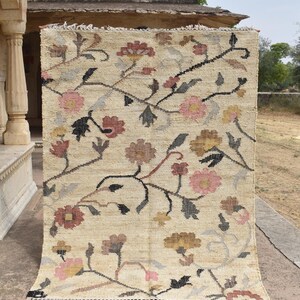 May include: A rectangular beige rug with a floral pattern. The rug features a repeating design of flowers in shades of pink, brown, and yellow, with black and gray stems and leaves. The rug has a fringed edge.