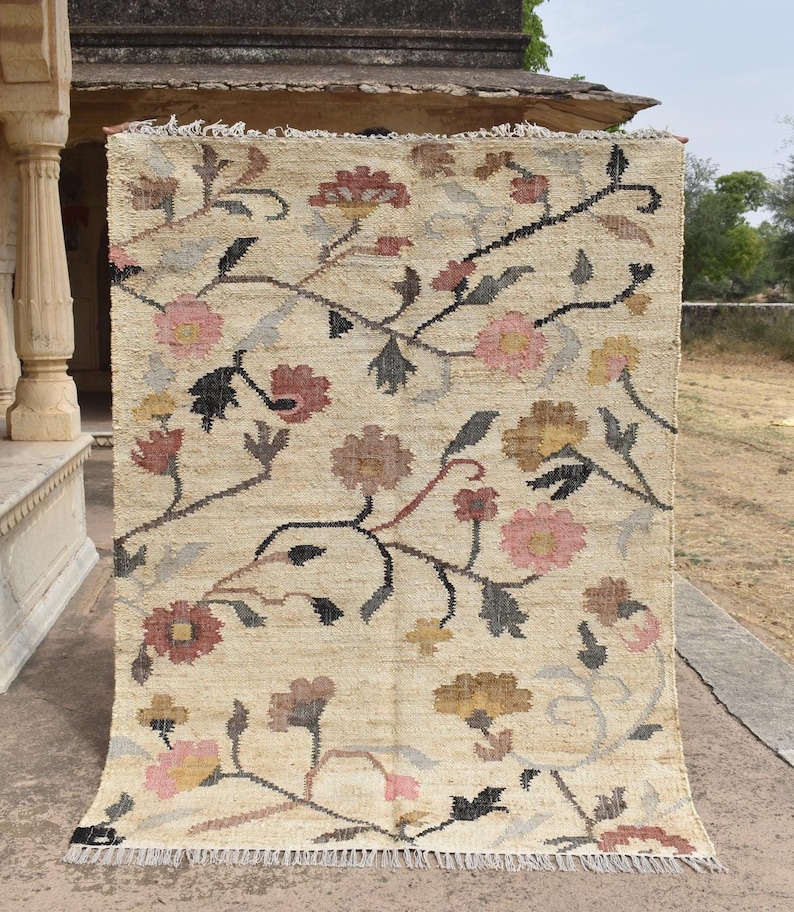 May include: A rectangular beige rug with a floral pattern. The rug features a repeating design of flowers in shades of pink, brown, and yellow, with black and gray stems and leaves. The rug has a fringed edge.