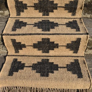 Op de afbeelding: Een vloerkleed van natuurlijke vezels met een geometrisch patroon in zwart en beige. Het vloerkleed heeft een franje rand en is geweven met een eenvoudig, rustiek ontwerp.