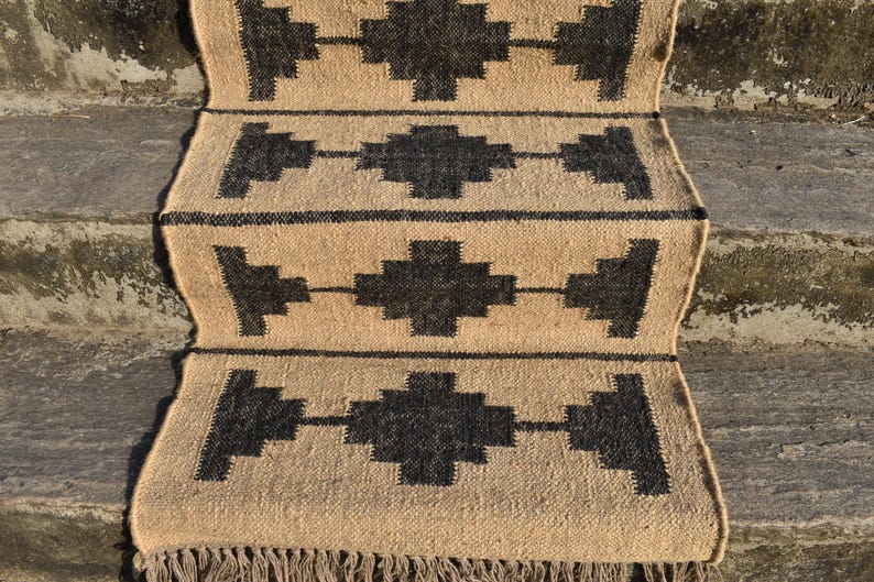 Op de afbeelding: Een vloerkleed van natuurlijke vezels met een geometrisch patroon in zwart en beige. Het vloerkleed heeft een franje rand en is geweven met een eenvoudig, rustiek ontwerp.