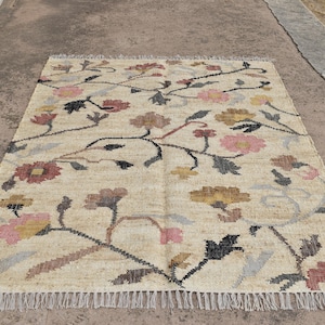 May include: Rectangular beige rug with a floral pattern. The rug features a repeating design of flowers in shades of pink, yellow, and brown, connected by dark stems. The rug has a fringed edge and is suitable for home decor.