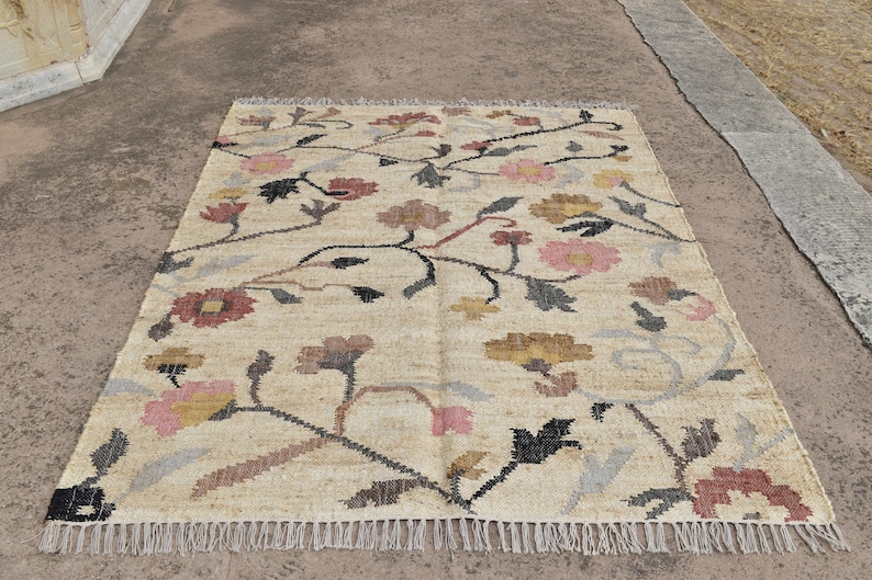 May include: Rectangular beige rug with a floral pattern. The rug features a repeating design of flowers in shades of pink, yellow, and brown, connected by dark stems. The rug has a fringed edge and is suitable for home decor.