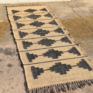 Op de afbeelding: Een vloerkleed van natuurlijke vezels met een geometrisch patroon in zwart en beige. Het vloerkleed heeft een franje rand.