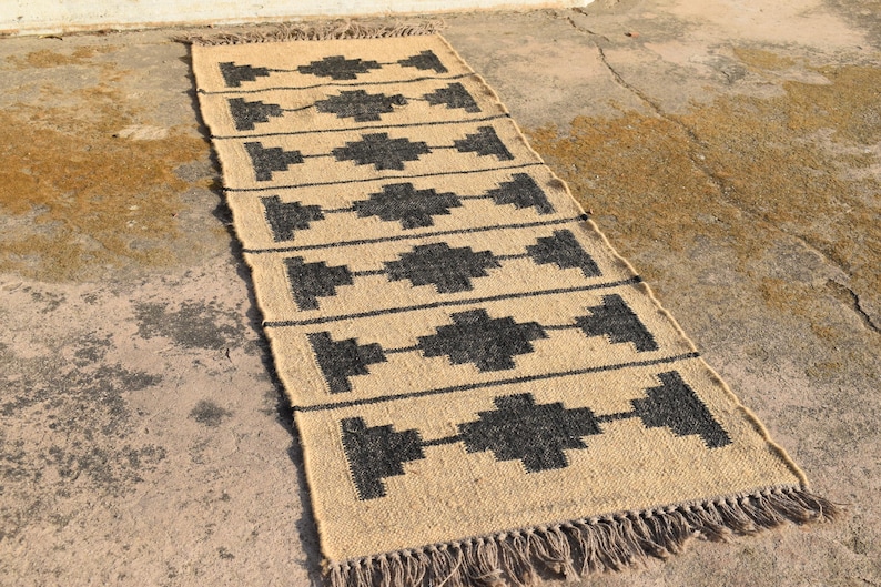 Op de afbeelding: Een vloerkleed van natuurlijke vezels met een geometrisch patroon in zwart en beige. Het vloerkleed heeft een franje rand.
