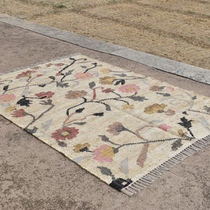May include: Rectangular beige rug with a floral pattern in pink, yellow, and brown. The rug has a fringe on the edges and is made of a woven material. The floral design features stems and leaves.