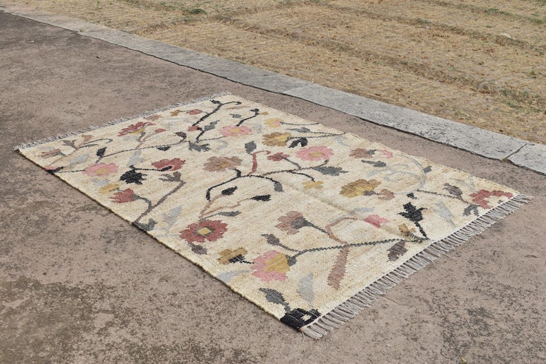 May include: Rectangular beige rug with a floral pattern in pink, yellow, and brown. The rug has a fringe on the edges and is made of a woven material. The floral design features stems and leaves.
