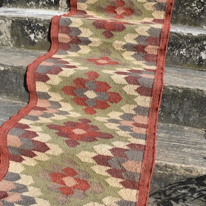 Puede incluir: Una alfombra de pasillo tejida con un estampado floral en tonos rojos, verdes, beige y grises. La alfombra tiene un borde rojo y se exhibe sobre escalones de hormigón. El diseño de la alfombra presenta motivos florales repetidos.
