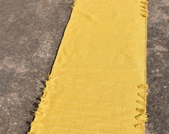 Op de afbeelding: Een rechthoekig, geel geweven vloerkleed met een getextureerd oppervlak en franjes langs de randen. Het vloerkleed is gemaakt van een dik, duurzaam materiaal en is geschikt voor binnen of buiten.