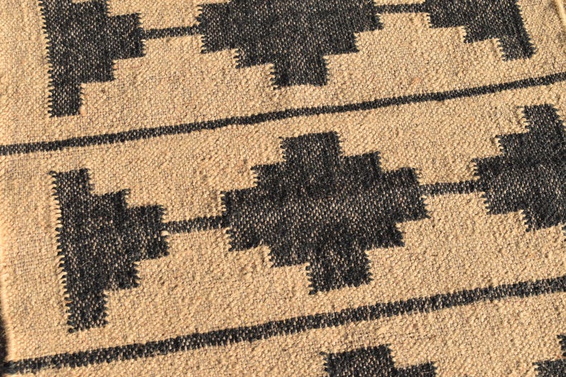 Op de afbeelding: Een close-up van een geweven tapijt met een geometrisch patroon. Het tapijt is gemaakt van natuurlijke vezels en heeft een beige achtergrond met zwarte geometrische vormen.