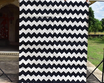 May include: A large, rectangular black and white chevron patterned rug. The rug is made of a woven material and is hanging from a metal rod. The rug has a fringe along the bottom edge. The background includes a building and greenery.