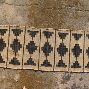 Op de afbeelding: Een geometrisch tapijt met zwart en beige patroon met franjes aan de uiteinden. Het tapijt heeft een eenvoudig ontwerp met herhalende zwarte vierkanten en rechthoeken op een beige achtergrond.