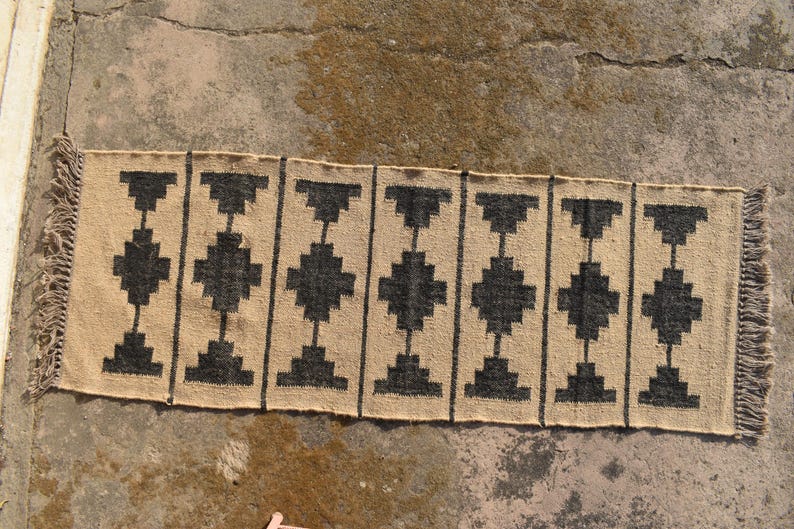Op de afbeelding: Een geometrisch tapijt met zwart en beige patroon met franjes aan de uiteinden. Het tapijt heeft een eenvoudig ontwerp met herhalende zwarte vierkanten en rechthoeken op een beige achtergrond.