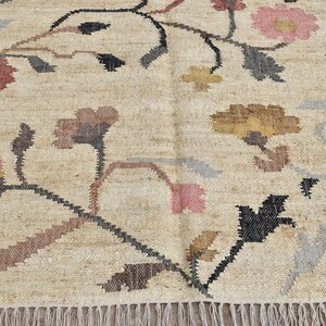 May include: Beige rug with floral design. The rug features a repeating pattern of flowers in shades of pink, brown, yellow, and gray, with black stems and leaves. The rug has a fringed edge.
